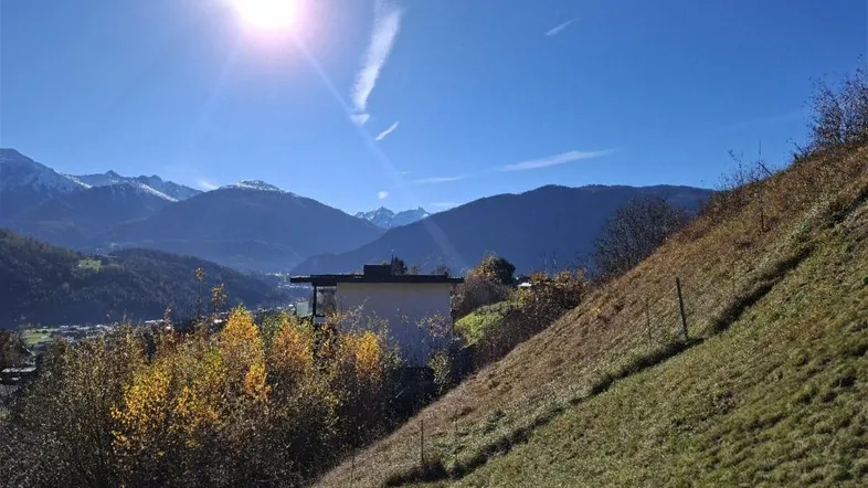 Sonnige Berglandschaft mit einem Haus am Hang und weitem Blick auf die Alpen.