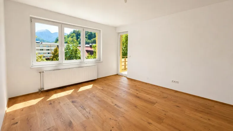 Helles Zimmer mit Holzboden, großen Fenstern und Zugang zum Balkon mit Bergblick.