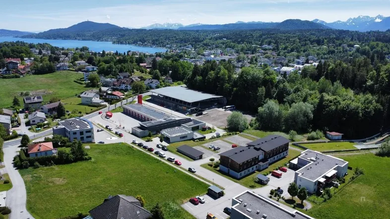 Weitläufige Luftaufnahme einer ländlichen Gegend mit einem See, Bergen im Hintergrund und verschiedenen Gebäuden inmitten grüner Natur.