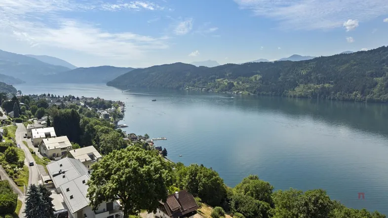 Panoramablick auf einen malerischen See, umgeben von grünen Hügeln und modernen Wohnhäusern am Ufer.