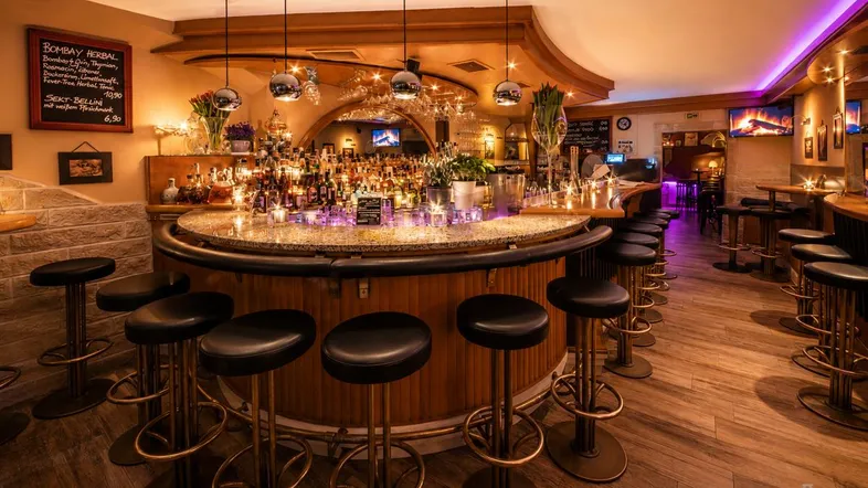 Eine elegant beleuchtete Bar mit einer großen, runden Theke, vielen Flaschen und bequemen Barhockern.