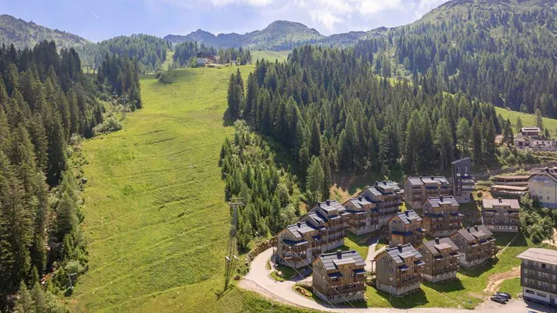 Luftaufnahme einer Ferienanlage mit mehreren Gebäuden inmitten einer grünen Berglandschaft und Skipiste.