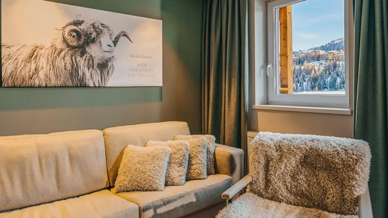 Gemütliches Wohnzimmer mit beigem Sofa, flauschigem Sessel und Fensterblick auf die verschneite Berglandschaft.