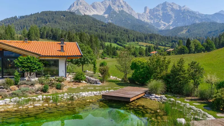Haus mit Teich und Holzsteg vor beeindruckender Bergkulisse unter blauem Himmel.