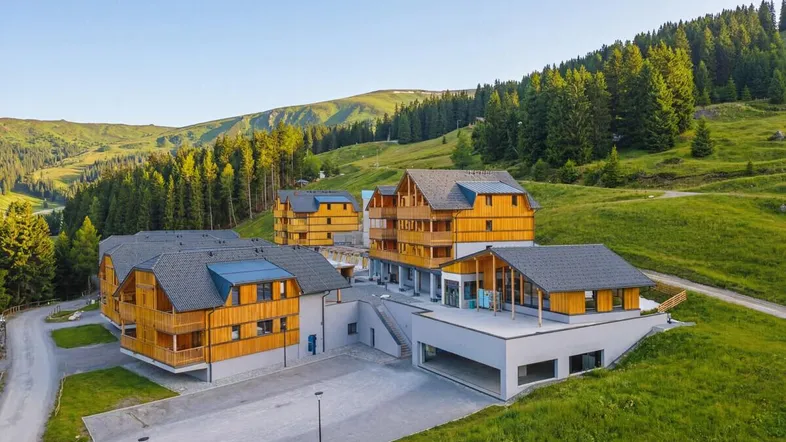 Luftaufnahme einer modernen Wohnanlage im alpinen Stil, umgeben von grünen Hügeln und Wäldern.
