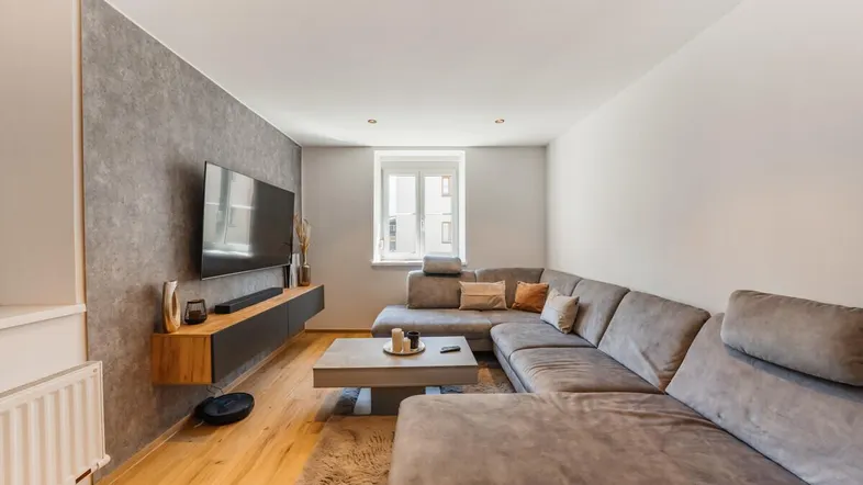 Modernes Wohnzimmer mit großem Ecksofa, Holzboden und einer Akzentwand in Betonoptik.
