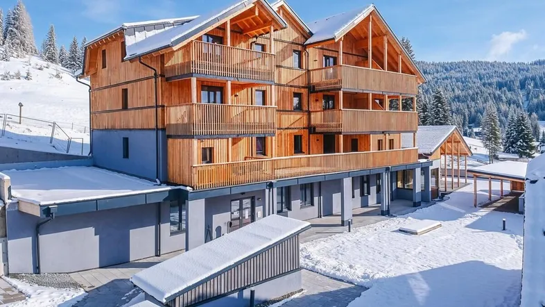 Modernes Holzgebäude mit mehreren Balkonen, umgeben von verschneiter Berglandschaft und Wäldern.