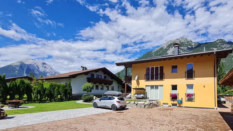 Modernes Haus mit gelber Fassade, gepflasterter Einfahrt und Bergblick unter blauem Himmel.