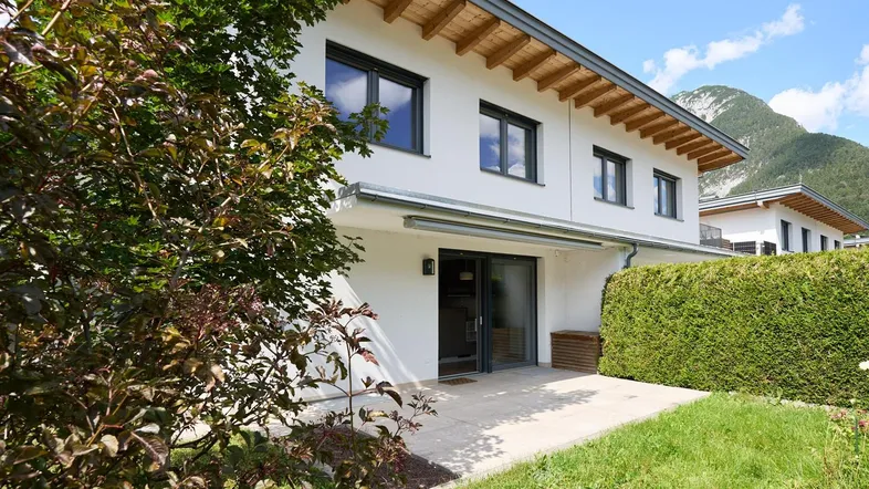 Moderne Hausfassade mit Terrasse, Rasenfläche und Bergblick unter blauem Himmel.