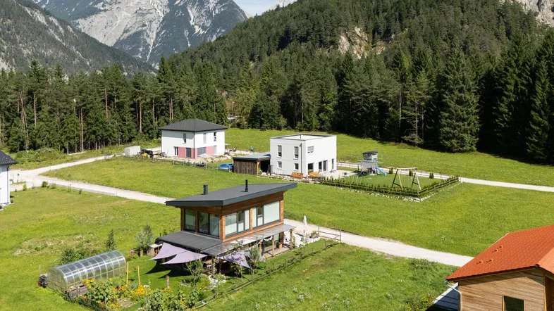 Luftaufnahme von modernen Häusern in einer grünen Tallandschaft mit Bergen und Wald im Hintergrund.