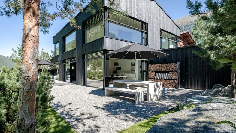 Modernes Haus mit dunkler Holzfassade, großer Terrasse und Bergblick in sonniger Umgebung.