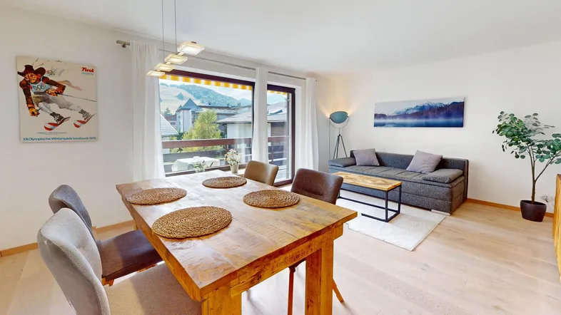 Helles Wohn- und Esszimmer mit Holztisch, bequemer Couch und Zugang zum Balkon mit Bergblick.