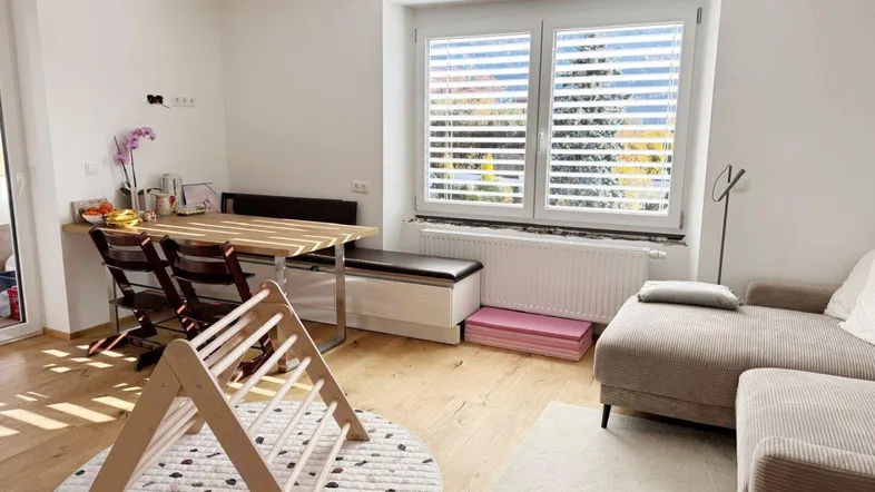 Heller Wohn- und Essbereich mit Holzboden, bequemer Couch und einem Fenster mit Jalousien.
