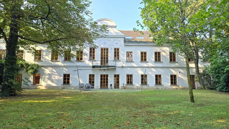 Großes weißes Gebäude mit vielen Fenstern und einem gepflegten Rasen, umgeben von hohen Bäumen.