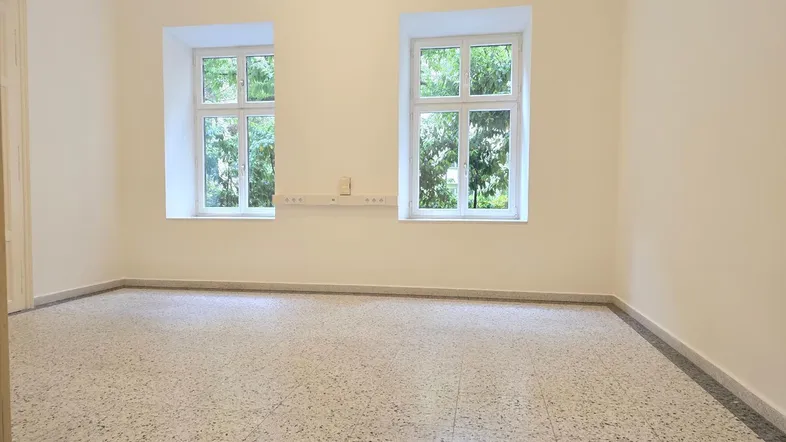 Leerer Raum mit zwei Fenstern, hellen Wänden und gesprenkeltem Terrazzoboden.