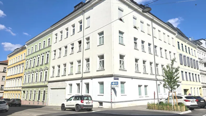 Weißes Mehrfamilienhaus mit mehreren Stockwerken und klassischen Fenstern an einer Straßenecke.