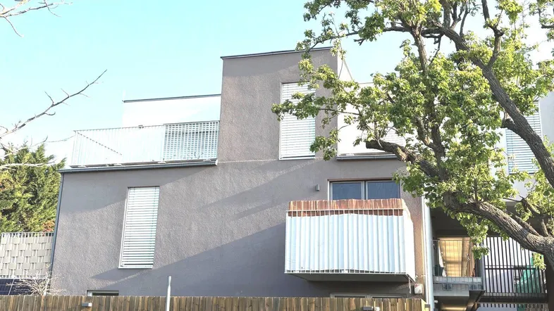 Moderne Fassade eines Wohngebäudes mit Balkon und Rollläden, umgeben von Bäumen.