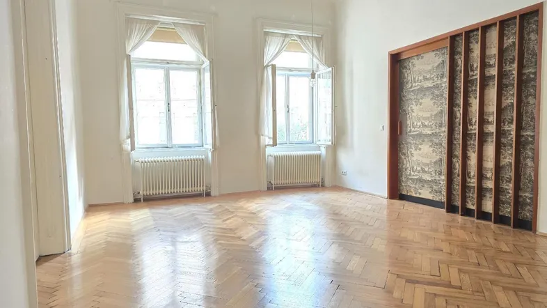Helles, leeres Zimmer mit Fischgrätparkett und zwei großen Fenstern, die viel Tageslicht hereinlassen.