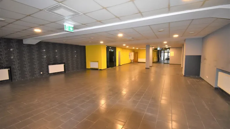 Großer, leerer Gewerberaum mit dunklem Fliesenboden, abgehängter Decke und gelben Akzentwänden.