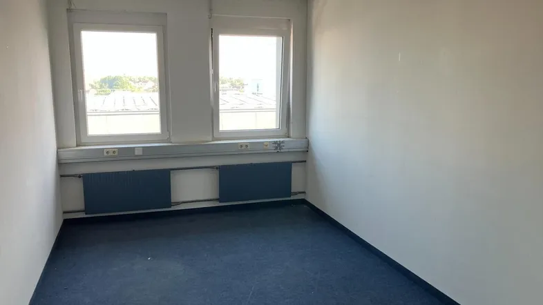 Leeres Büro mit zwei Fenstern, blauem Teppichboden und weißen Wänden, ausgestattet mit Heizkörpern.