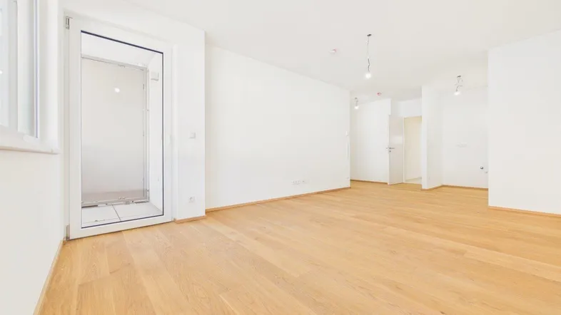 Helles, leeres Zimmer mit Holzboden und Zugang zu einem Balkon oder einer Terrasse.