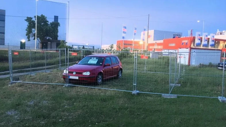 Roter PKW auf einem grasbewachsenen Grundstück, umzäunt von temporären Metallzäunen, mit Gewerbegebäuden im Hintergrund.