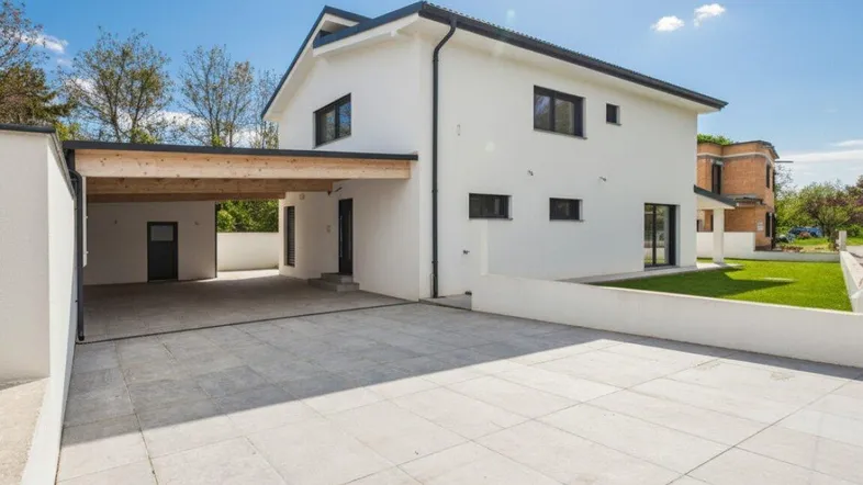Modernes Einfamilienhaus mit großem gepflasterten Vorplatz und Carport unter strahlend blauem Himmel.