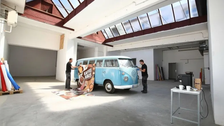 Großer, heller Innenraum mit Oberlichtern, in dem ein VW-Bus für eine Veranstaltung vorbereitet wird.