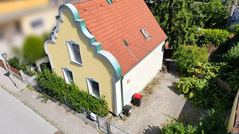 Historisches Haus mit gelber Fassade und rotem Ziegeldach in grüner Umgebung.