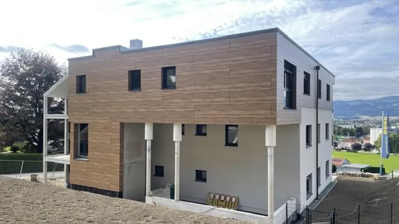 Moderne Fassade eines Neubaus mit Holzverkleidung und weißem Putz, umgeben von Baustellengelände.