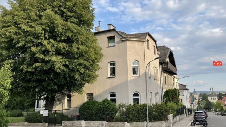 Ansicht eines Mehrfamilienhauses an einer Straßenecke mit altem Baumbestand und gepflegter Hecke.