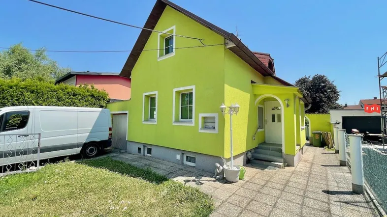 Gelbgrünes Einfamilienhaus mit Satteldach, gepflastertem Vorplatz und einem weißen Lieferwagen davor.
