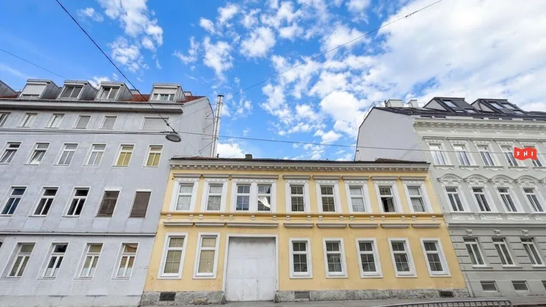 Gelbes Gebäude mit vielen Fenstern und einem großen Tor an einer städtischen Straße, umgeben von anderen Bauten.