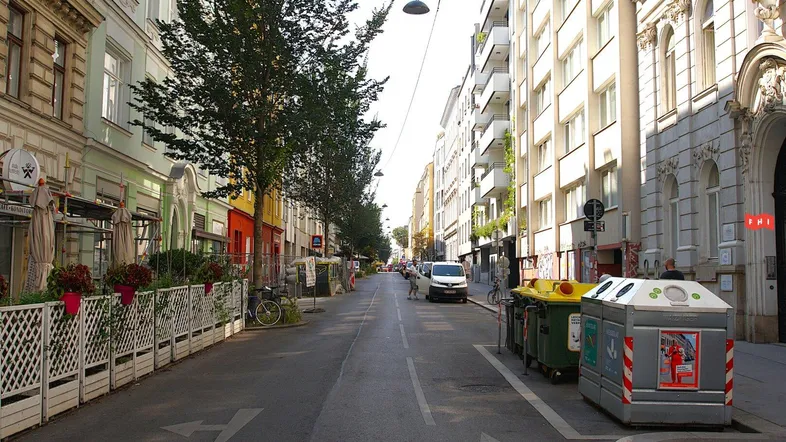 Eine belebte Straße mit Gebäuden, Bäumen und Müllcontainern, die das städtische Umfeld zeigt.