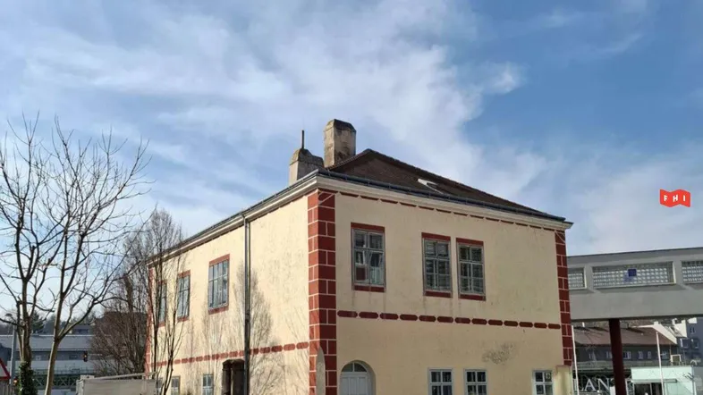 Historisches Gebäude mit beiger Fassade und roten Zierelementen unter blauem Himmel, umgeben von kahlen Bäumen.