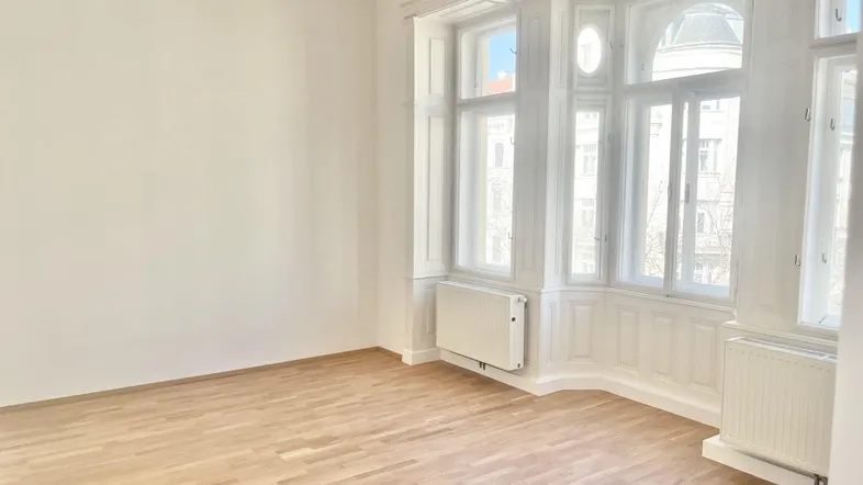 Helles Wohnzimmer mit elegantem Parkettboden, hohen Decken und einem großen Erkerfenster.