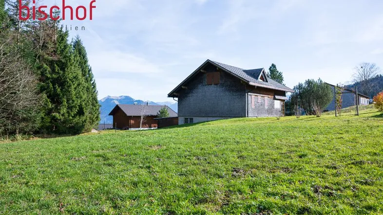 Landhaus mit Holzfassade und Satteldach, umgeben von grünen Wiesen und Bäumen mit Bergblick.