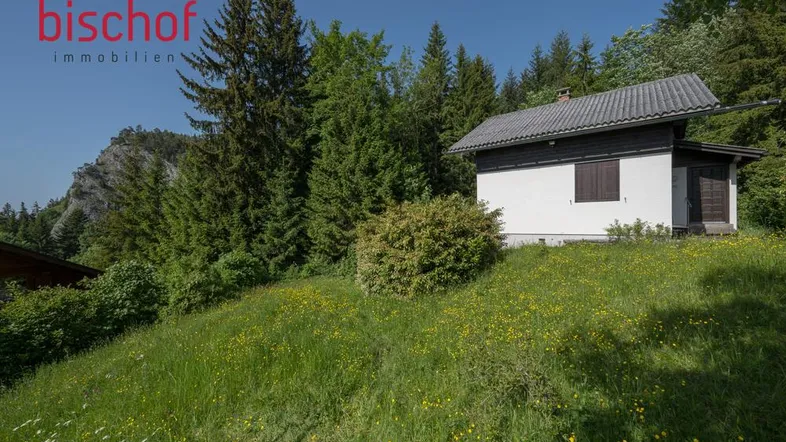 Kleines weißes Gebäude mit dunklem Dach, umgeben von grünen Wiesen und Bäumen vor einer Felswand.