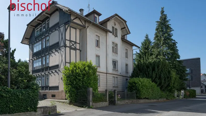 Historisches Gebäude mit Fachwerkdetails und viel Grün in sonniger Umgebung.