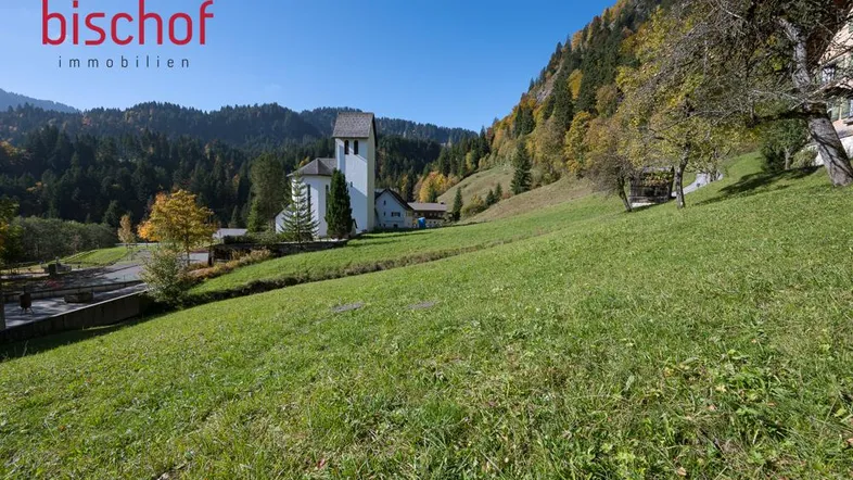 Weitläufige grüne Wiese mit Blick auf eine Kirche und die umliegende Berglandschaft unter blauem Himmel.
