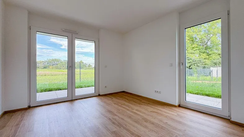 Heller Raum mit Laminatboden und großen Fenstern, die Zugang zu einer Terrasse und Blick ins Grüne bieten.