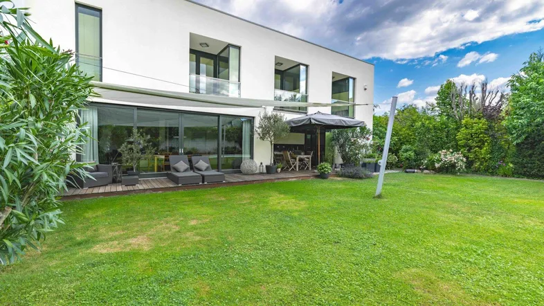 Moderner Gartenbereich mit großer Rasenfläche, Holzterrasse und Liegestühlen hinter dem Haus.