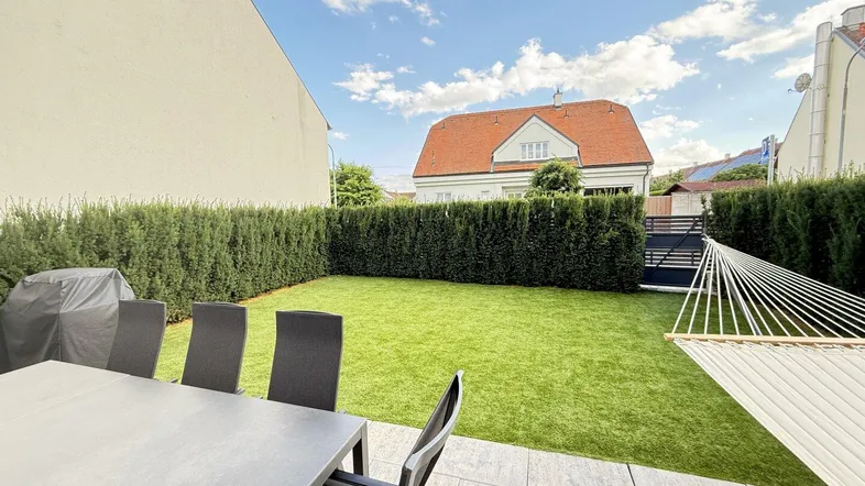 Gepflegter Gartenbereich mit Rasenfläche, Terrasse und Gartenmöbeln, umgeben von einer hohen Hecke.