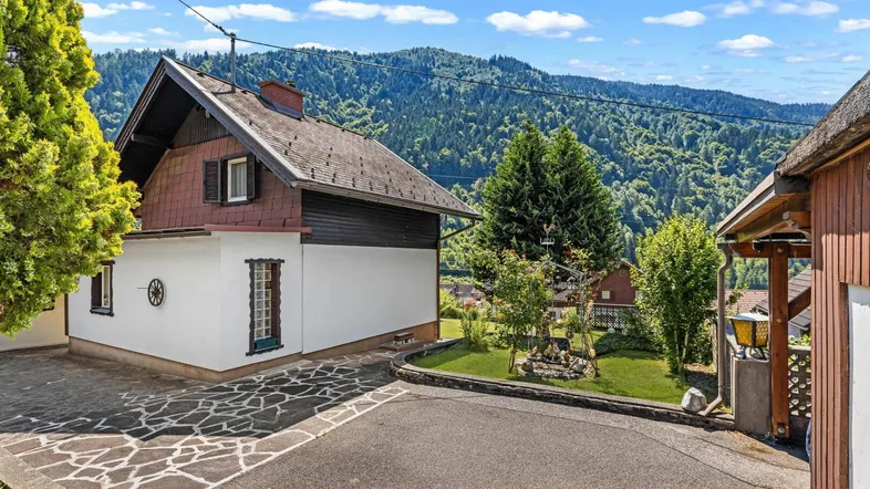 Hausfassade mit gepflastertem Vorplatz und Blick auf die umliegenden Berge und Wälder.