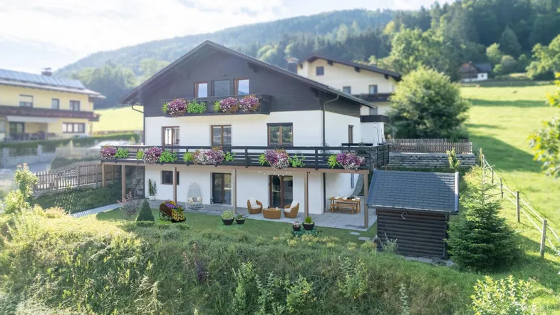 Haus mit Balkonen, Terrasse und Garten in bergiger Landschaft mit viel Grün.