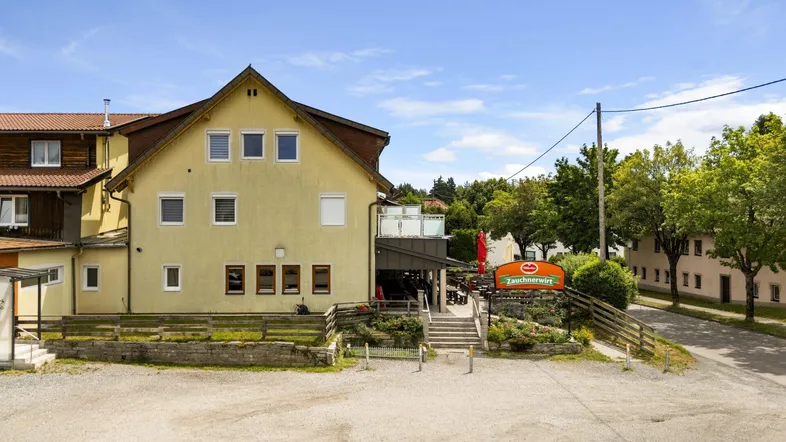 Das Gebäude präsentiert sich als Gasthof mit gelber Fassade und einem Außenbereich, umgeben von Bäumen unter blauem Himmel.
