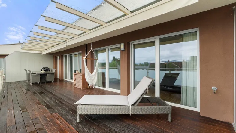 Große Dachterrasse mit Holzdeck, Sonnenliege und Essbereich unter einer Glasüberdachung.