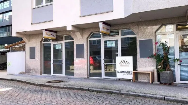 Die Außenansicht des Geschäftsgebäudes zeigt große Fensterfronten und einen gepflasterten Gehweg mit einem Werbeschild.