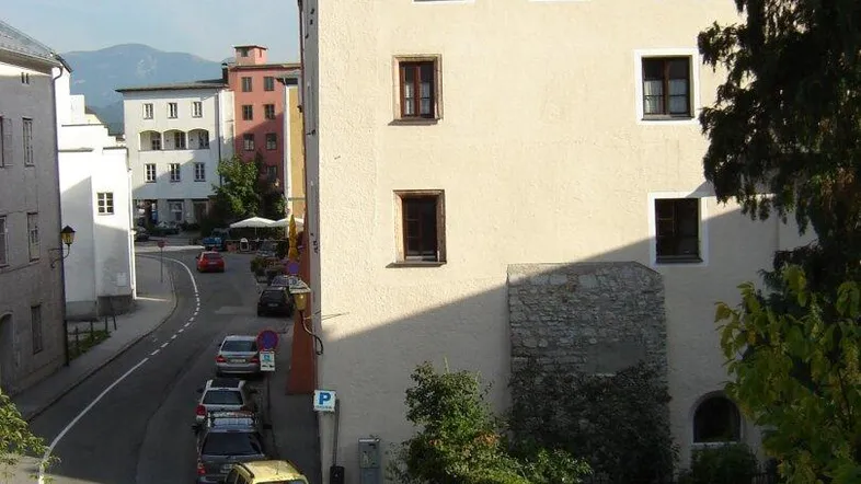 Gebäudeansicht an einer Straße mit geparkten Autos und Bergen im Hintergrund.