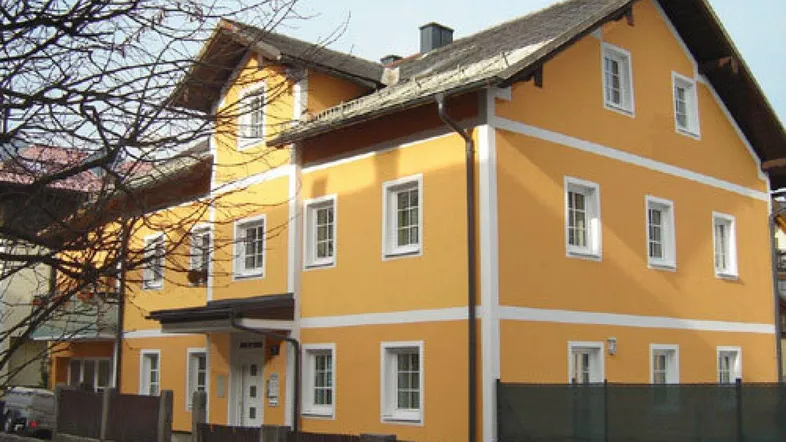 Gelbe Fassade eines Mehrfamilienhauses mit weißen Fensterrahmen und Satteldach in gepflegtem Zustand.